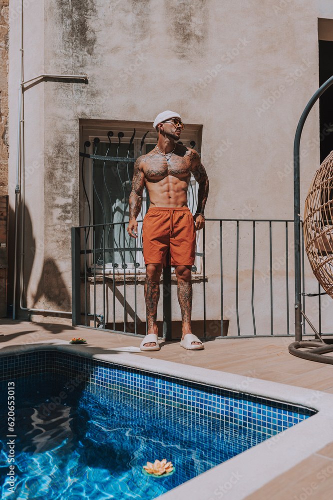 CHico joven musculado con tatuajes posando sin camiseta en piscina con estilo de hotel en día soleado 