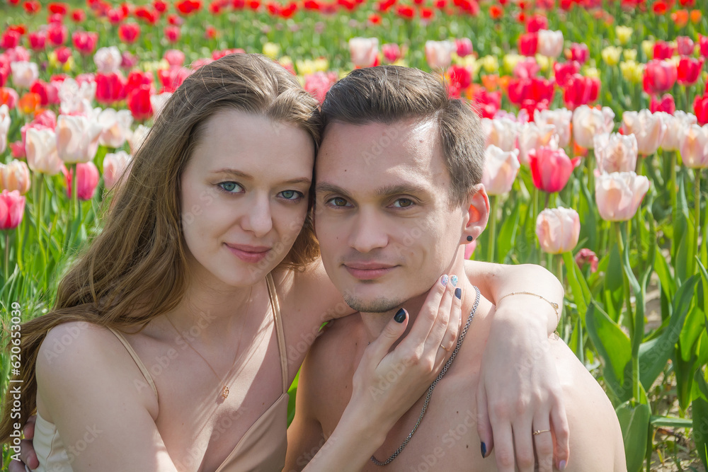 Young beautiful smiling couple sits on the background of a field of colorful tulips.