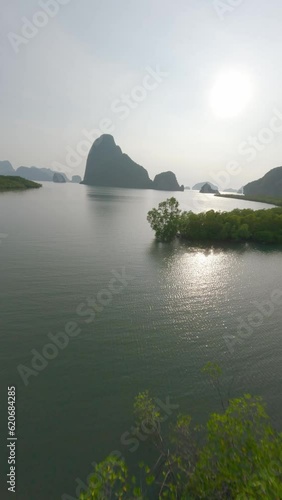 Vertical video. Aerial view forward flying lush sunny green trees exotic forest vegetation sea mountain rock island. FPV sport drone shot greenery wood plant scenery natural rock cliff cloudy sky sun