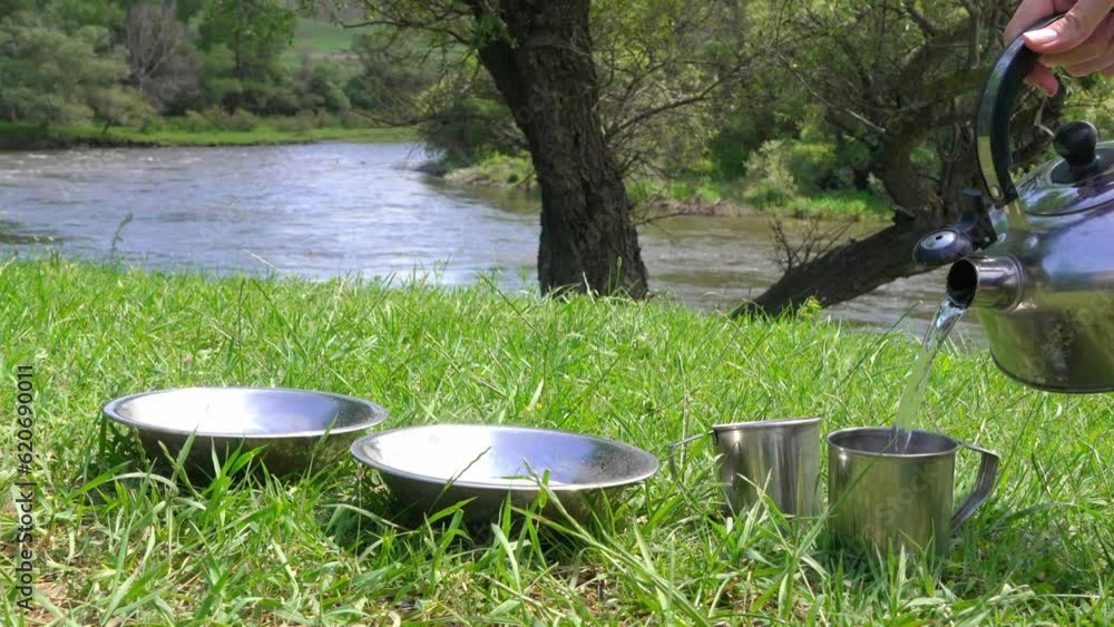 Picnic on banks river against background green grass. Woman's hand pours water from kettle into iron camping bowls and mugs, tea or coffee and oatmeal are quickly prepared. Breakfast on trip or hike. Picnic on banks river against background green grass. Woman's hand pours water from kettle into iron camping bowls and mugs, tea or coffee and oatmeal are quickly prepared. Breakfast on trip or hike.