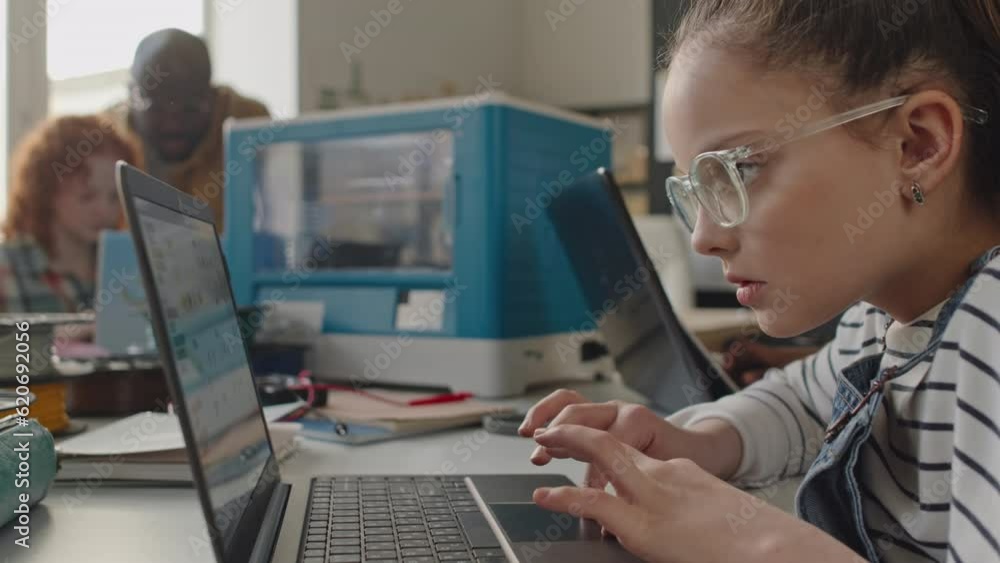 Tilt down shot of smart little girl in eyeglasses using laptop at desk while learning 3D printing on school lesson Tilt down shot of smart little girl in eyeglasses using laptop at desk while learning 3D printing on school lesson
