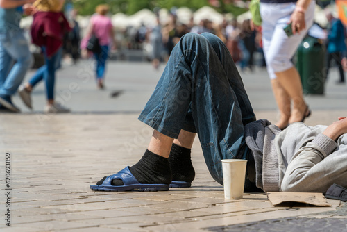 Authentic tramp sleeping on a cobbled street on a sunny summer day with a paper cup for donations against the background of passers-by and tourists. Global connectivity, diversity and inclusion