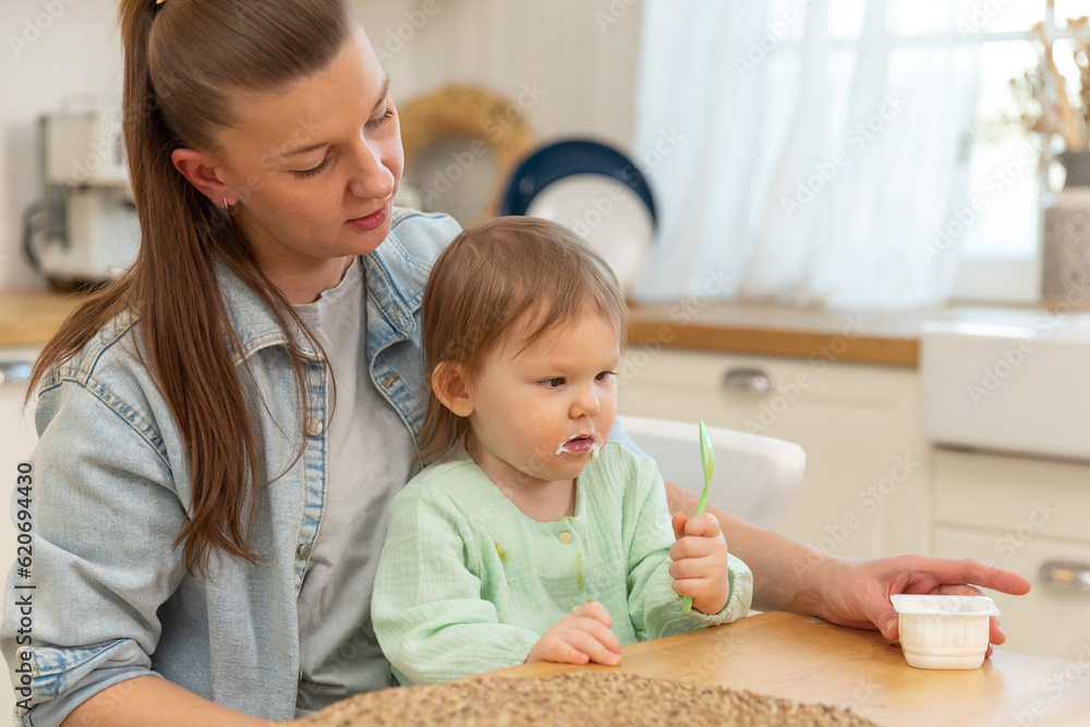 Happy family at home. Mother feeding her baby girl from spoon in kitchen. Little toddler child with messy funny face eats healthy food at home. Young woman mom giving food to kid daughter Happy family at home. Mother feeding her baby girl from spoon in kitchen. Little toddler child with messy funny face eats healthy food at home. Young woman mom giving food to kid daughter