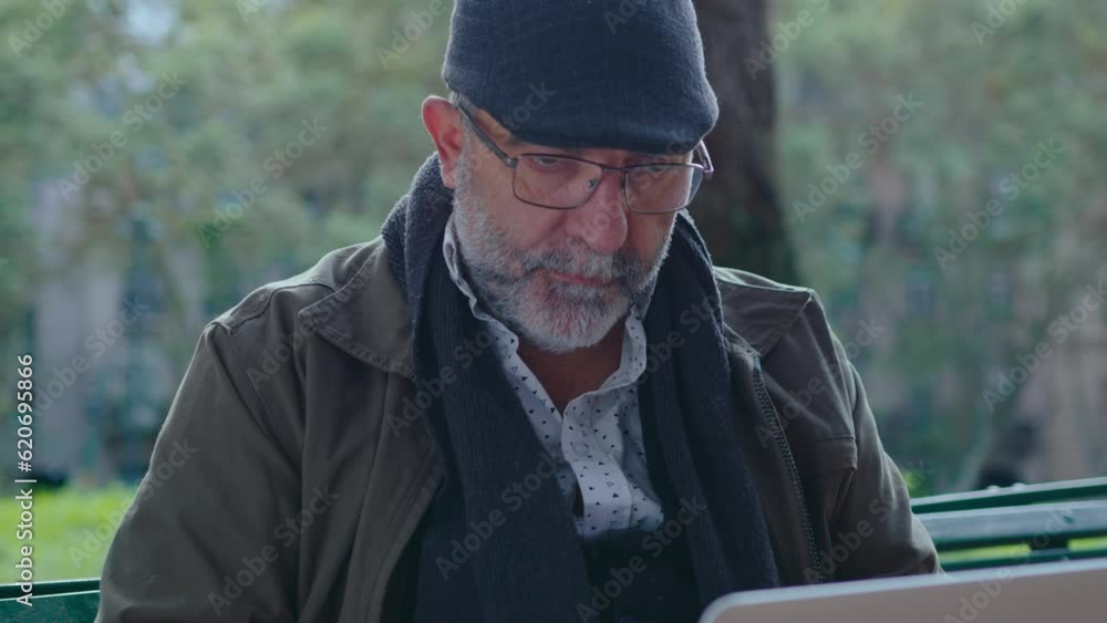 Senior man with gray beard wearing eyeglasses and cap sitting on bench in the park, chin stroking, thinking thoughtfully and using laptop outdoors Senior man with gray beard wearing eyeglasses and cap sitting on bench in the park, chin stroking, thinking thoughtfully and using laptop outdoors