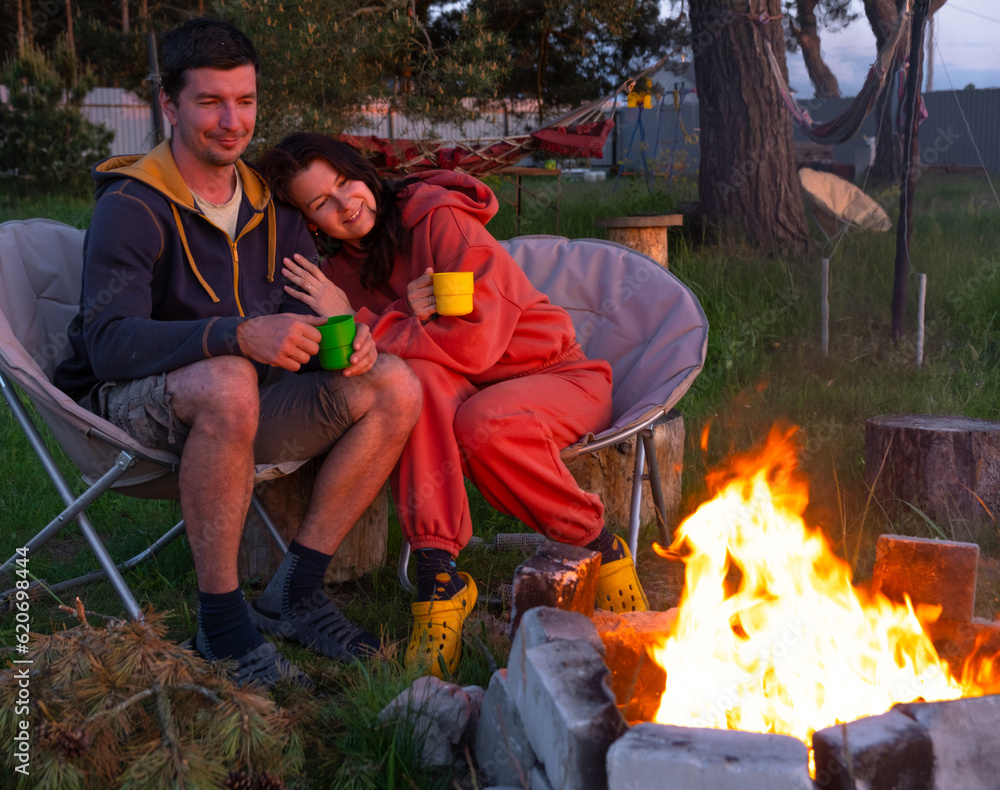 Couple in love is sitting by the outdoor campfire in the courtyard of the house on camping chairs, a romantic evening. a man and a woman warm their hands by the fire, drink tea Couple in love is sitting by the outdoor campfire in the courtyard of the house on camping chairs, a romantic evening. a man and a woman warm their hands by the fire, drink tea