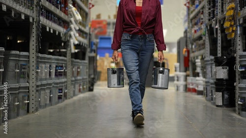 Woman shown from waist down, carrying to cans of paint walking down paint aisle in hardware store