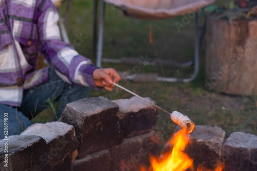 A girl in a plaid shirt roasts marshmallows on a fire in the yard of the house. Evening family get-together by the campfire, outdoor tourist chairs