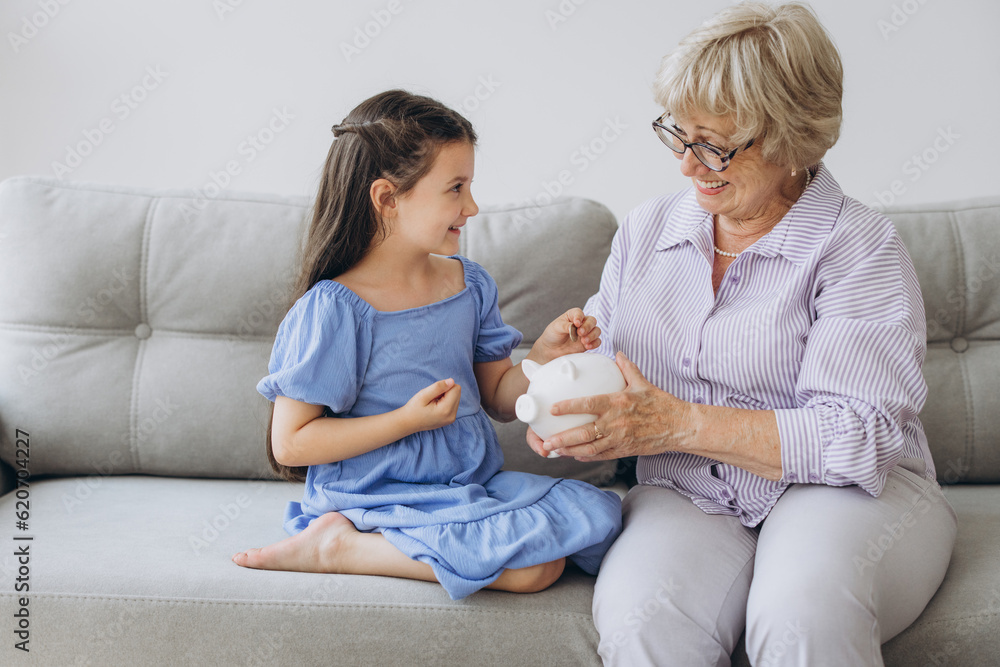 Grandmother and her granddaughter Putting Coin Money In Piggybank At Home. Personal Savings, Bank Safety And Financial Investments Concept Grandmother and her granddaughter Putting Coin Money In Piggybank At Home. Personal Savings, Bank Safety And Financial Investments Concept