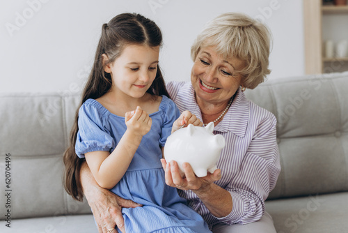 Grandmother and her granddaughter Putting Coin Money In Piggybank At Home. Personal Savings, Bank Safety And Financial Investments Concept