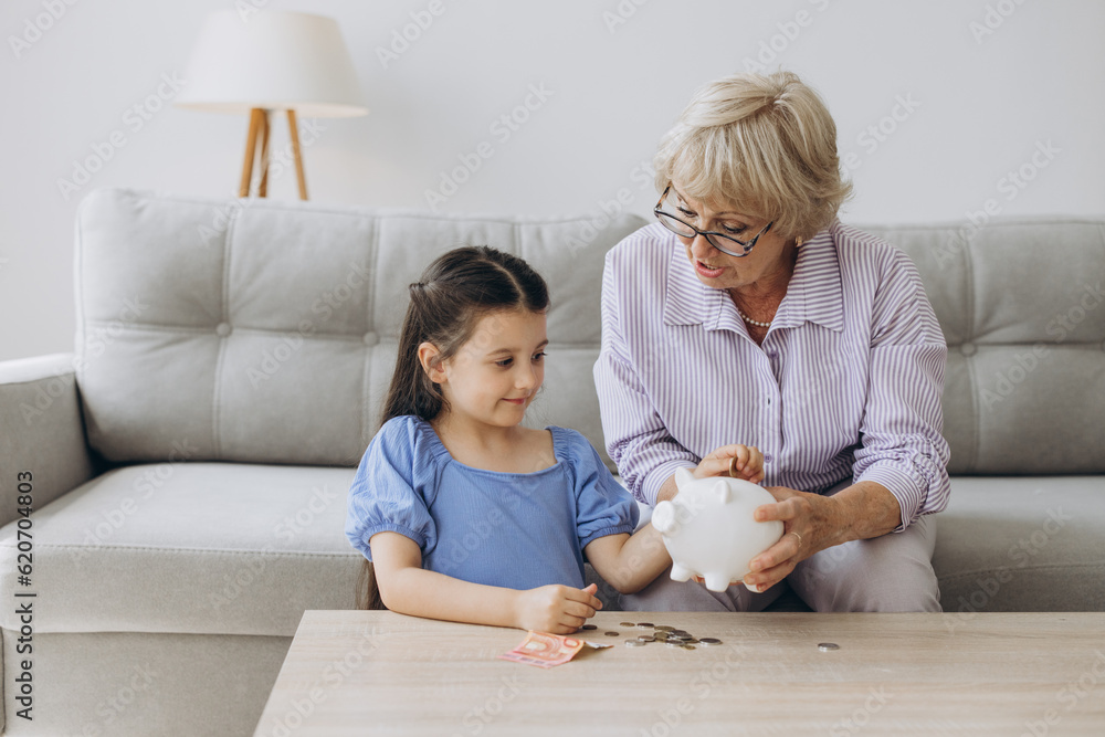 Grandmother and her granddaughter Putting Coin Money In Piggybank At Home. Personal Savings, Bank Safety And Financial Investments Concept Grandmother and her granddaughter Putting Coin Money In Piggybank At Home. Personal Savings, Bank Safety And Financial Investments Concept