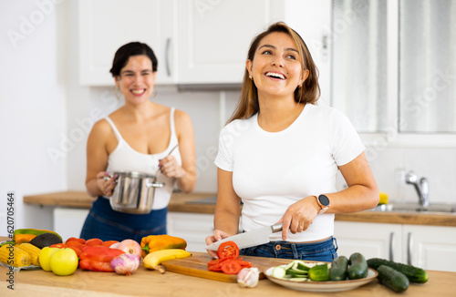 Smiling young Hispanic woman cooking with sister in home kitchen, preparing fresh vegetable salad. Happy family relationship concept