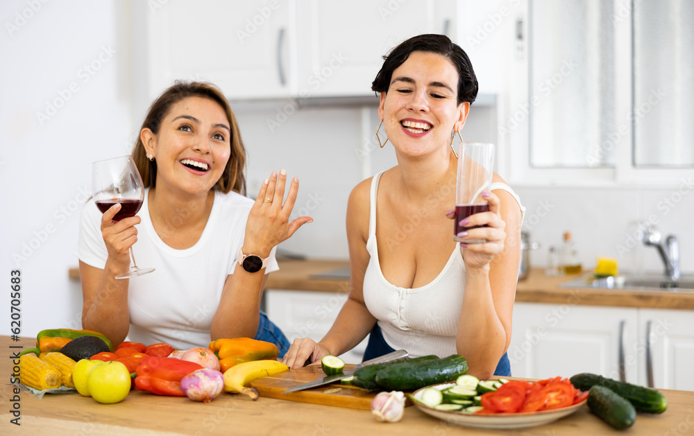 Two happy female friends preparing salad and drinking red wine together in modern kitchen