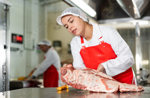 Focused young butcher shop saleswoman cutting fresh raw pork ribs at meat processing table..