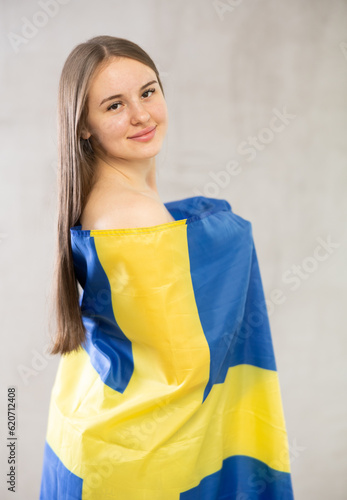 Girl holding large Sweden flag in hands. Young teenage girl draped over shoulders, wrapped herself in fabric of national Swedish flag. Studio shot, gray background