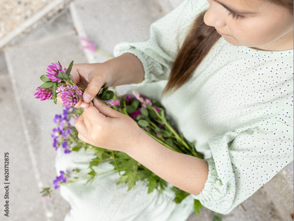 A girl in a green dress sits on the steps and weaves a wreath of collected wildflowers, top view