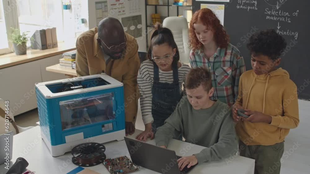 High angle shot of school boy surrounded by classmates and teacher sitting at desk, using laptop and 3D printer during additive manufacturing lessonHigh angle shot of school boy surrounded by classmat High angle shot of school boy surrounded by classmates and teacher sitting at desk, using laptop and 3D printer during additive manufacturing lessonHigh angle shot of school boy surrounded by classmat