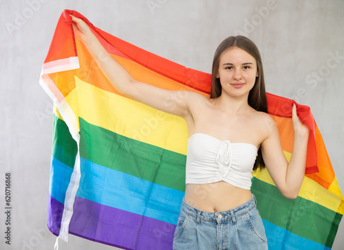 Positive young teen girl in casual clothes holds unfurled striped colorful flag of LGBT in hands raised above head against gray wall  studio shot. Studio shot  isolated