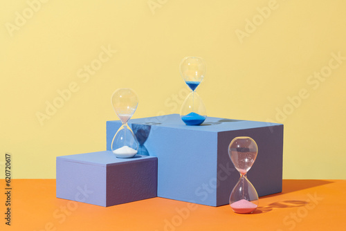 three hourglasses with color sand standing on blue podium photo