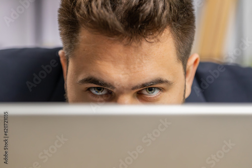 Close-up confident businessman hiding behind laptop computer, looking at camera, spying his colleagues working, peeping. Professional freelancer man looking from behind computer, cunning eyes glance