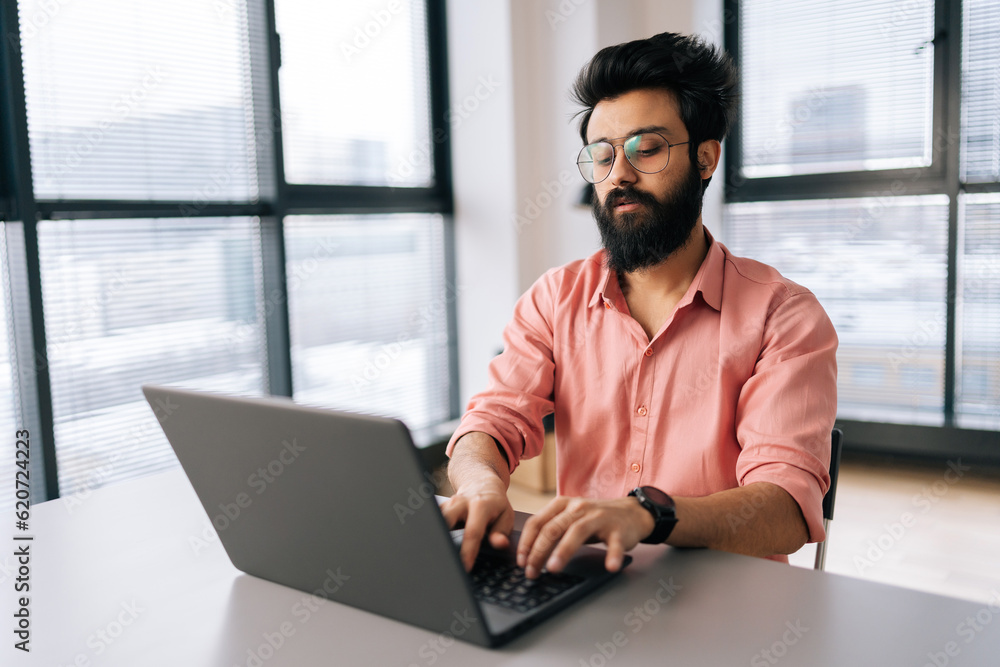 Fototapeta premium Focused Indian freelancer male in stylish eyeglasses working on laptop sitting at desk in light coworking office room by window. Bearded business man wearing casual clothes looking at computer screen.
