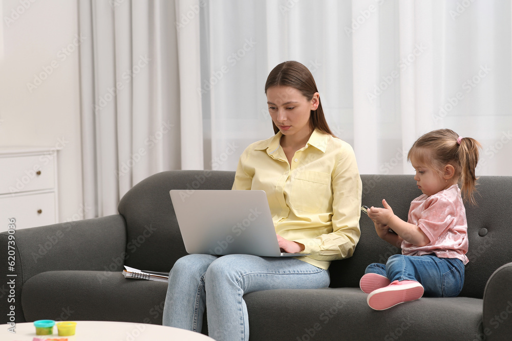 Mother working remotely on laptop while her daughter playing with smartphone at home Mother working remotely on laptop while her daughter playing with smartphone at home