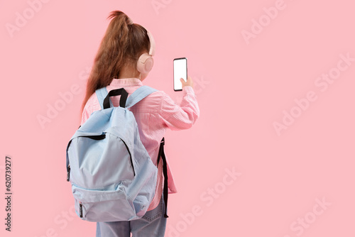 Little schoolgirl in headphones with backpack and mobile phone on pink background, back view