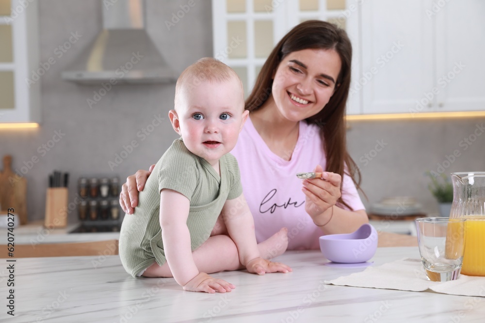 Fototapeta premium Happy young woman feeding her cute little baby at table in kitchen