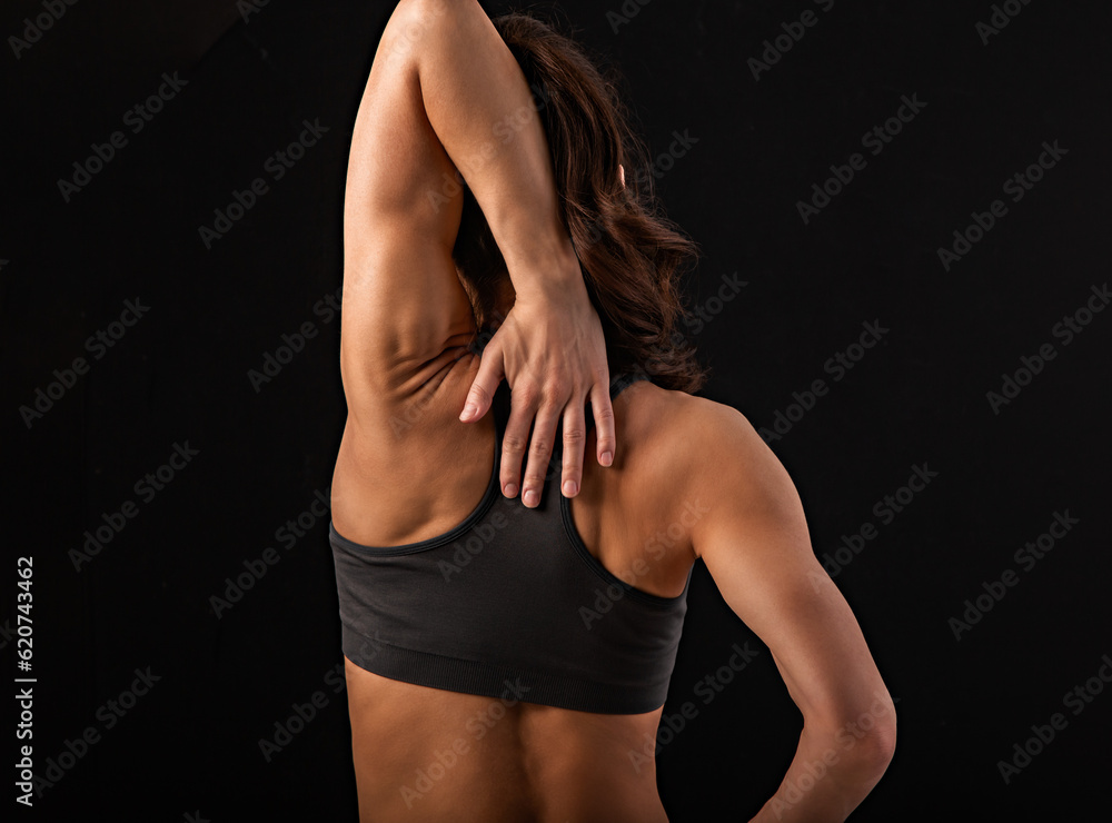Naklejka premium Female sporty muscular with ponytail doing stretching workout of the shoulders, blades in sport bra, holding the neck the hand on black studio background with empty copy space. Back view.