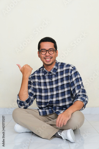 Adult Asian man sitting cross legged on the floor smiling and pointing to the right side photo