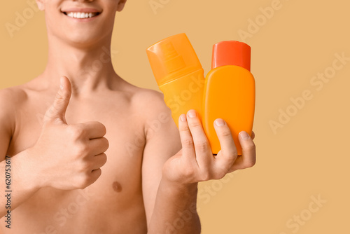 Happy young man with bottles of sunscreen cream showing thumb-up gesture on orange background