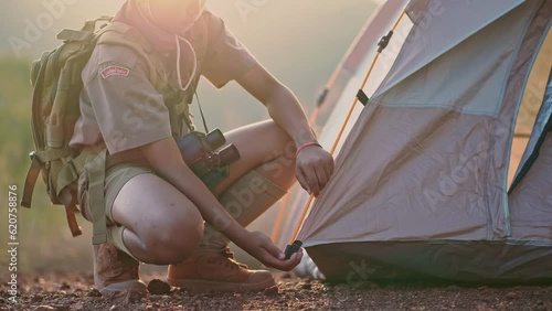 An Asian Boy Scout Sits on the Ground and Packs his Backpack in a Mountain Scout Camp near Sunset.