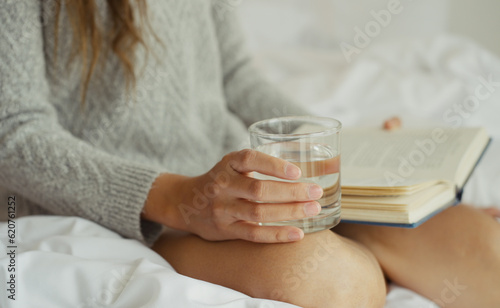 Young asian woman drinking water while reading a book sitting on the bed in the morning.