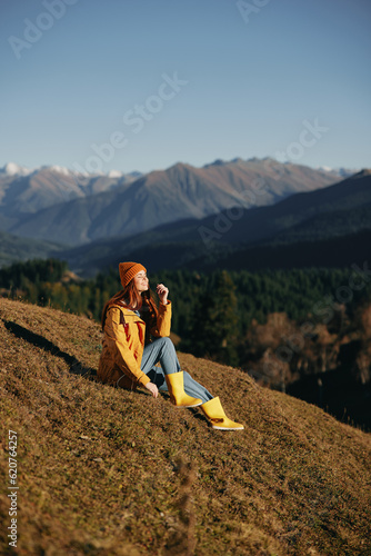 Woman full-length sitting resting on a hill and looking at the mountains in a yellow raincoat and jeans happy camping trip in the autumn, freedom lifestyle 