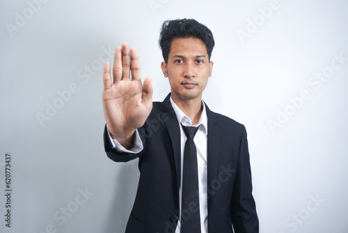 Serious Asian man gesturing STOP, showing palm to camera over grey studio background. Young man expressing his negative attitude, rejecting or prohibiting something