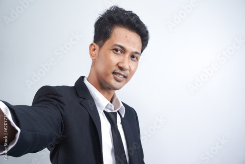 Handsome young man wearing suit taking a selfie by his phone, standing on a white background.