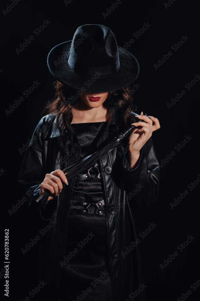 Sexy female dominant mistress with a whip for BDSM sex with submission and domination in a leather raincoat and a hat in the noir style Sexy female dominant mistress with a whip for BDSM sex with submission and domination in a leather raincoat and a hat in the noir style