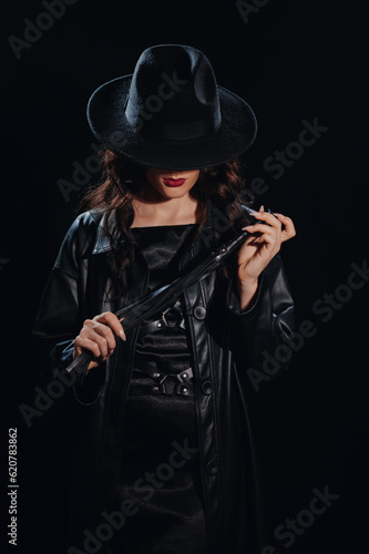 Sexy female dominant mistress with a whip for BDSM sex with submission and domination in a leather raincoat and a hat in the noir style