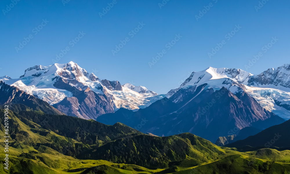 An awe-inspiring landscape featuring a series of towering mountains with snow-capped peaks, nestled amidst lush green valleys and surrounded by a clear blue sky.