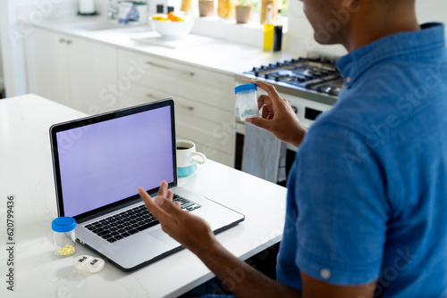 Biracial man holding medication making video call in kitchen using laptop with copy space on screen
