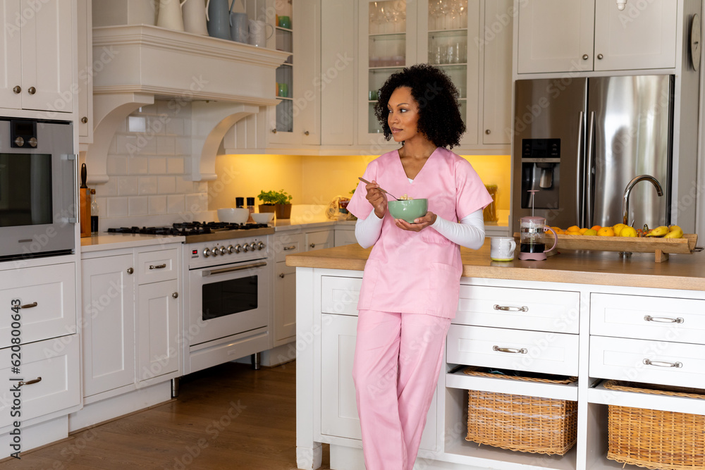 Happy biracial female medical worker in scrubs eating bowl of cereal in kitchen, copy space Happy biracial female medical worker in scrubs eating bowl of cereal in kitchen, copy space