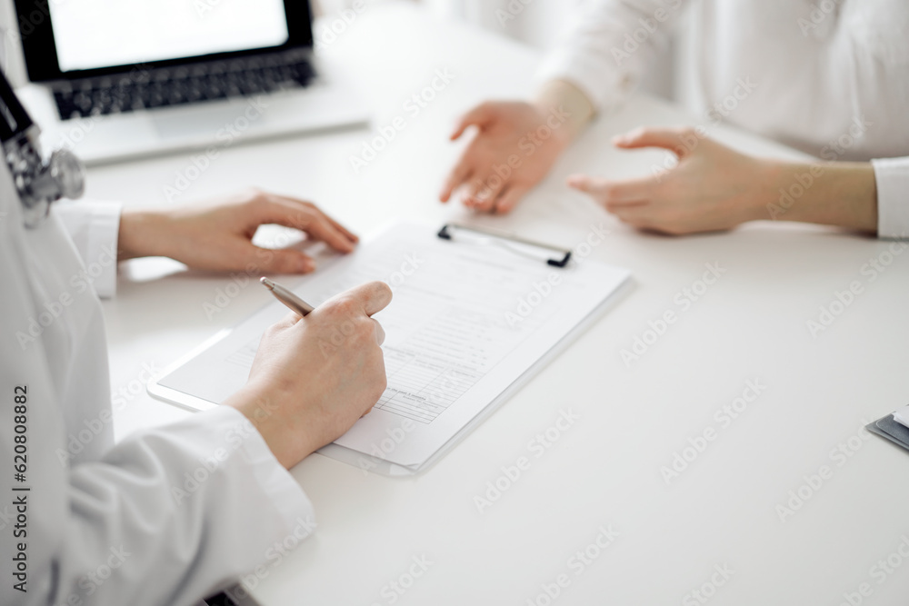 Doctor and patient sitting near each other at the white desk in clinic. Female physician is listening filling up a records form. Medicine concept Doctor and patient sitting near each other at the white desk in clinic. Female physician is listening filling up a records form. Medicine concept