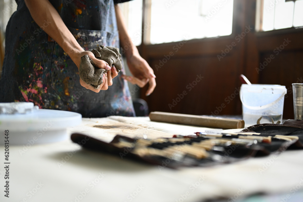 Obraz premium Pottery artist in apron kneading piece of raw clay with his hands on wooden table. Handicraft, creativity, hobby and activity concept