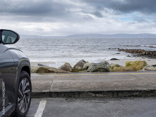 Dark color car parked by the ocean with view on a stunning ocean landscape with mountains in the background. Dramatic cloudy sky. Galway bay, Ireland, Travel and sightseeing.