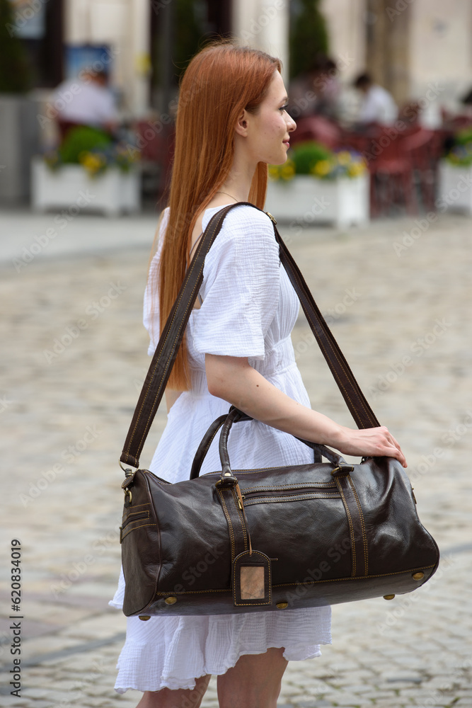 young beautiful redhair girl wearing white summer dress with brown travel bag posing on the streets young beautiful redhair girl wearing white summer dress with brown travel bag posing on the streets