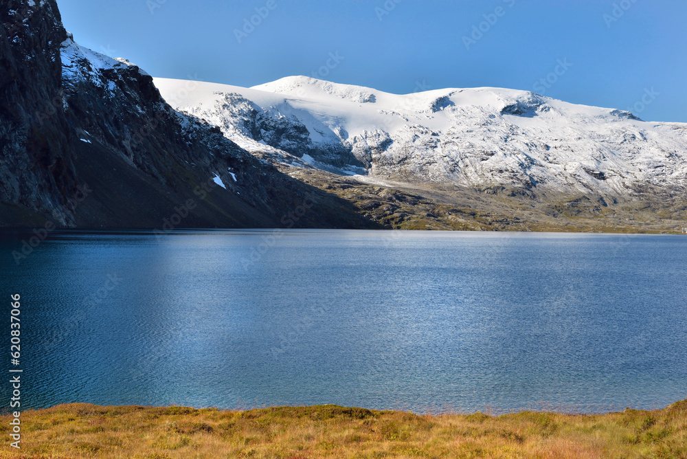 beautiful norway landscape with blue lake nestled in the snow-capped mountains and surrounded by autum-coloured vegetation under blue sky