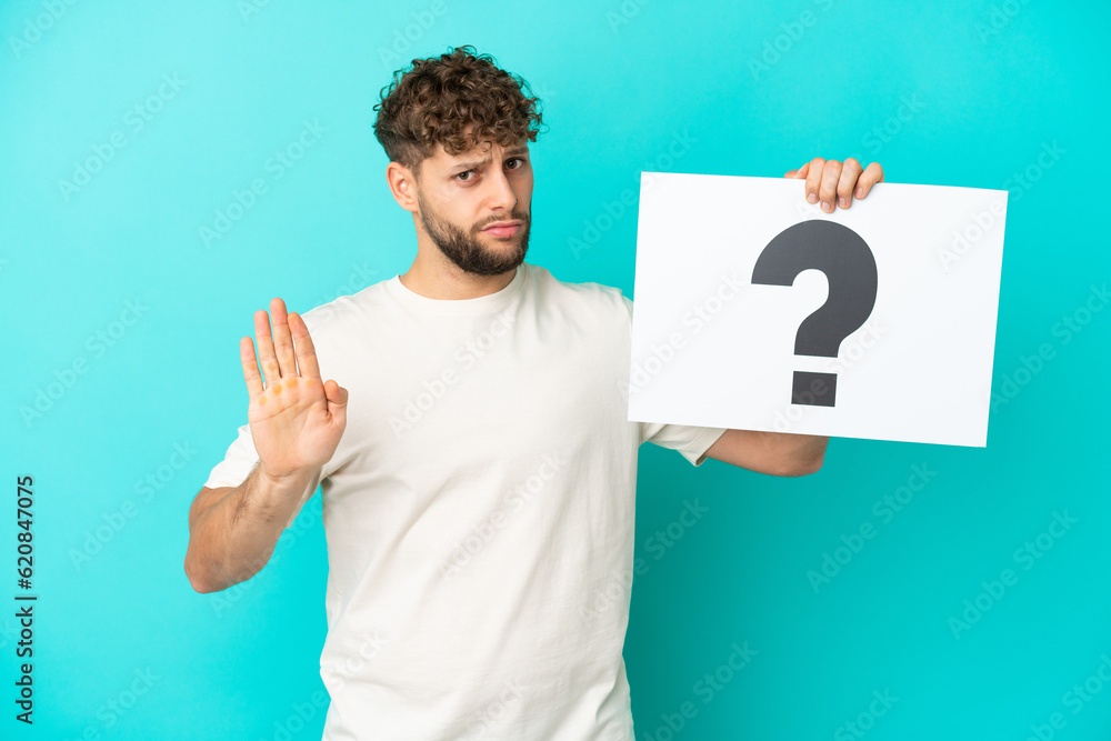 Young handsome caucasian man isolated on blue background holding a placard with question mark symbol and doing stop sign Young handsome caucasian man isolated on blue background holding a placard with question mark symbol and doing stop sign