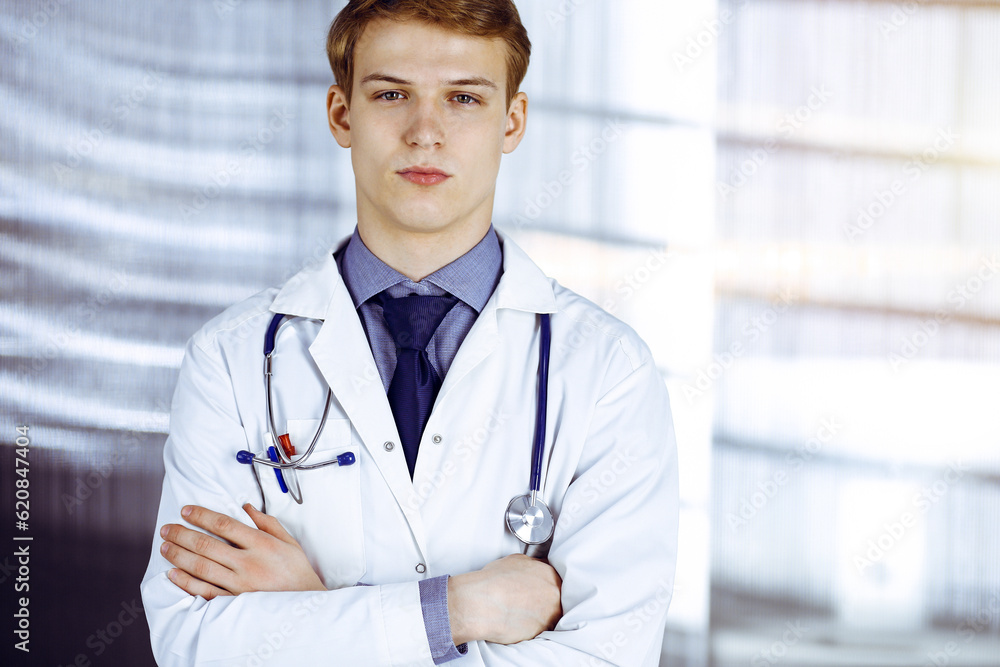 Young doctor standing with crossed arms in sunny clinic near his working place. Medicine concept Young doctor standing with crossed arms in sunny clinic near his working place. Medicine concept