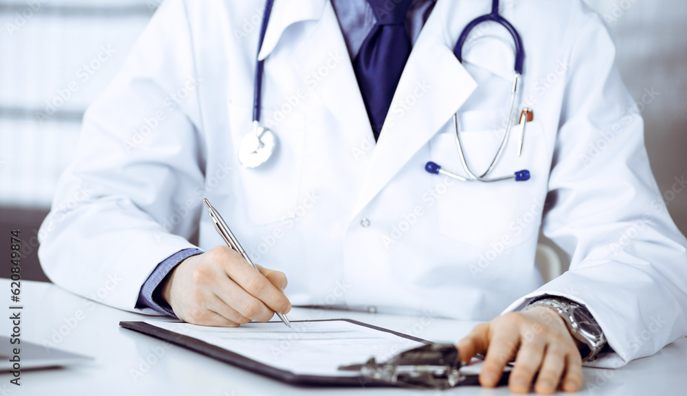 Unknown male doctor sitting and working with clipboard in clinic at his working place, close-up. Young physician at work. Perfect medical service, medicine concept Unknown male doctor sitting and working with clipboard in clinic at his working place, close-up. Young physician at work. Perfect medical service, medicine concept