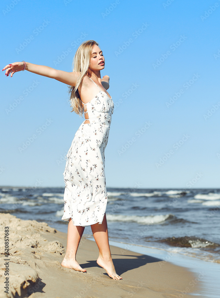 Happy blonde beautiful woman on the ocean beach standing in a white summer dress, open arms Happy blonde beautiful woman on the ocean beach standing in a white summer dress, open arms