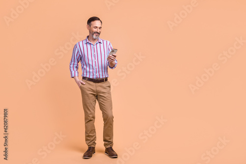 Full length photo of handsome positive person use smart phone typing empty space advert isolated on beige color background